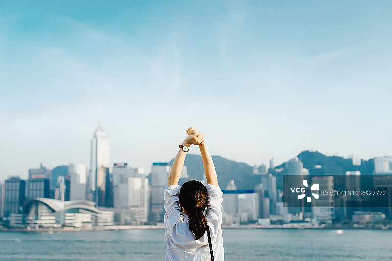 后视图的女性旅行者享受她的时间在香港，深呼吸与高举的手对维多利亚港和城市天际线图片素材