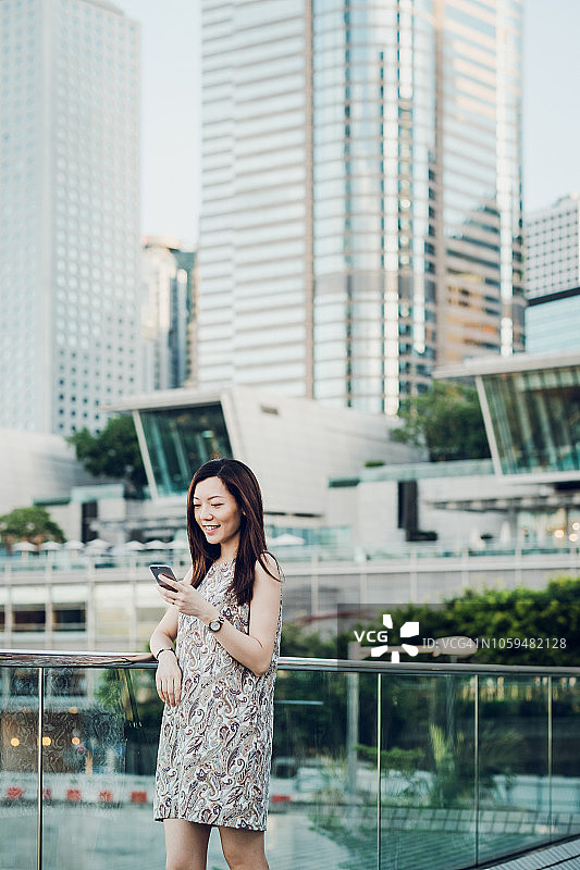 美丽微笑的亚洲女人在城市里使用手机，对着中央商务区的高楼大厦图片素材