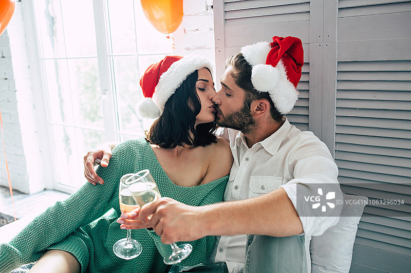 幸福的年轻夫妇戴着圣诞帽拥抱亲吻，在明亮的卧室里喝着香槟庆祝圣诞节图片素材