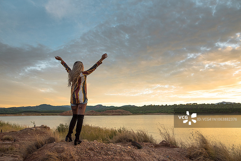 日落时分，一个年轻女子在湖边举起双手图片素材