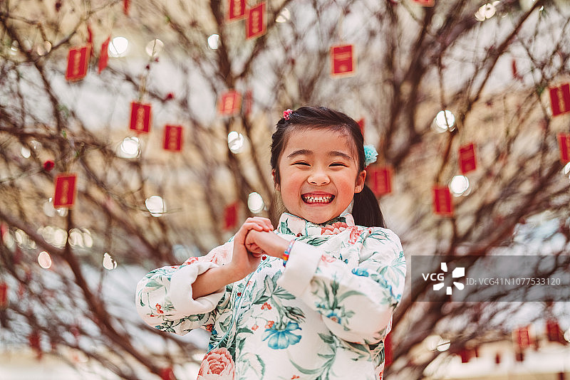 新年的时候，一个可爱的小女孩站在一棵桃花树前，对着镜头微笑着，用“恭喜发财”的手势庆祝新年图片素材
