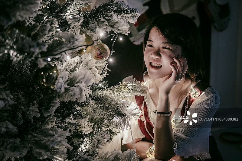 一位亚裔华人女性坐在圣诞树前庆祝圣诞节，微笑着看着它图片素材