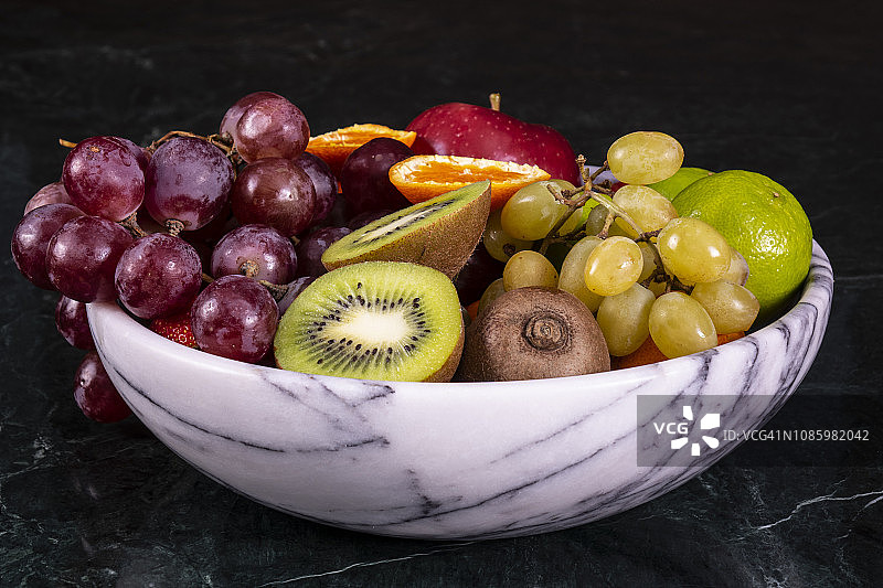 新鲜的热带水果在碗上黑色大理石背景。菠萝，木瓜，芒果，猕猴桃，葡萄，石榴，橘子，香蕉，苹果，西瓜片，火龙果图片素材