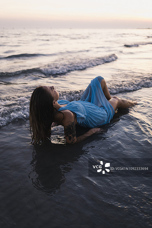 年轻的纹身女人穿着蓝色的衣服躺在海边的水日落图片素材