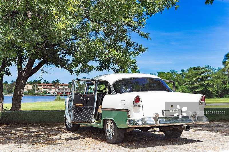 美国绿白色的经典汽车与开放的门停在树下的海滩在古巴巴拉德罗-古巴报道图片素材