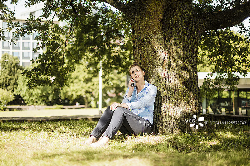 一个女人坐在城市公园里，靠在一棵树上打电话图片素材