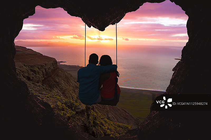 一对夫妇在秋千上凝视着海面上的日落，这是一个心形的浪漫风景。图片素材