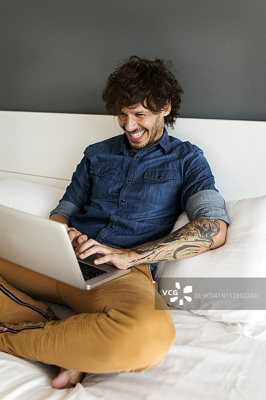 快乐的纹身男人坐在床上用笔记本电脑图片素材