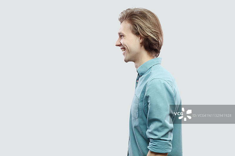 侧面的肖像快乐英俊的长头发金发青年男子在蓝色休闲衬衫站和看着与牙齿微笑。图片素材