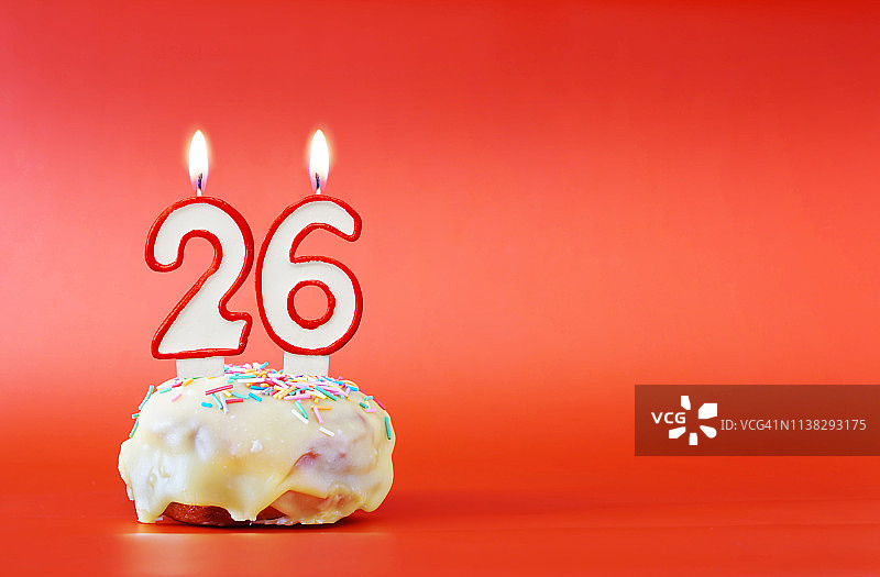二十六岁生日。纸杯蛋糕，上面有燃烧着的白色蜡烛，形状是26号。鲜艳的红色背景与复制空间图片素材