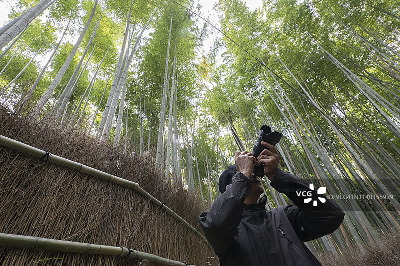 在日本京都的岚山竹林里，一名穿着黑色夹克的男子拿着相机。——图片图片素材