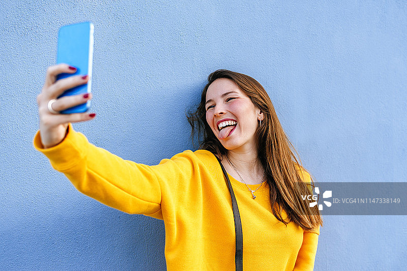 快乐的年轻女子在墙上自拍伸出舌头图片素材