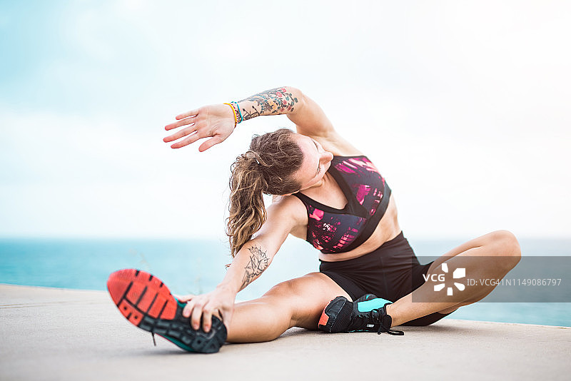 运动年轻的纹身女人伸展在海边。图片素材