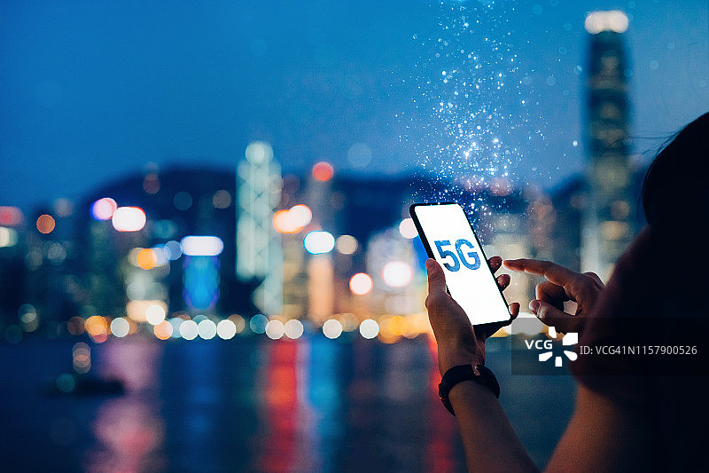 晚上，一名年轻女子在维多利亚港的海滨长廊上使用智能手机，背景是香港标志性的城市天际线，5G通信的概念被分解成轻粒子图片素材