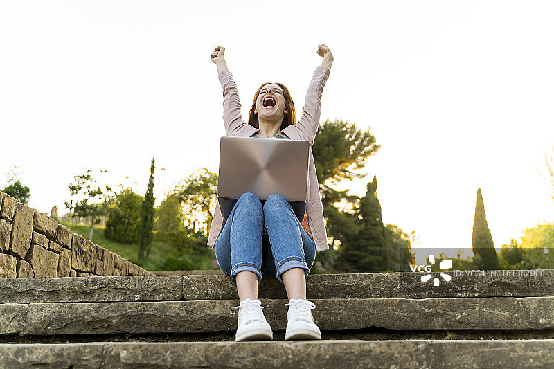 年轻的红发女子使用笔记本电脑，坐在公园的台阶上欢呼图片素材