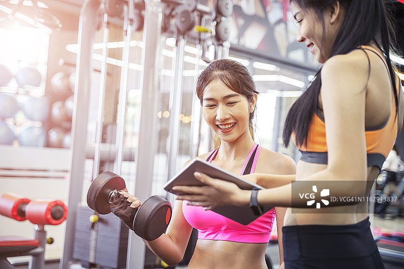 年轻的亚洲女子在健身房和她的私人教练锻炼。私人教练帮助妇女使用沉重的哑铃。图片素材