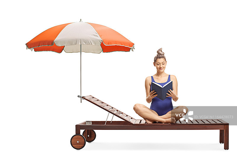一名年轻女子穿着游泳衣坐在伞下的日光浴床上看书图片素材