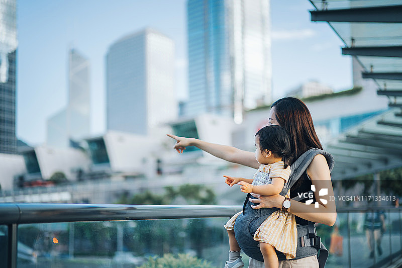 年轻的亚洲母亲指着她的小女儿，在市中心的城市露台上对着现代摩天大楼图片素材