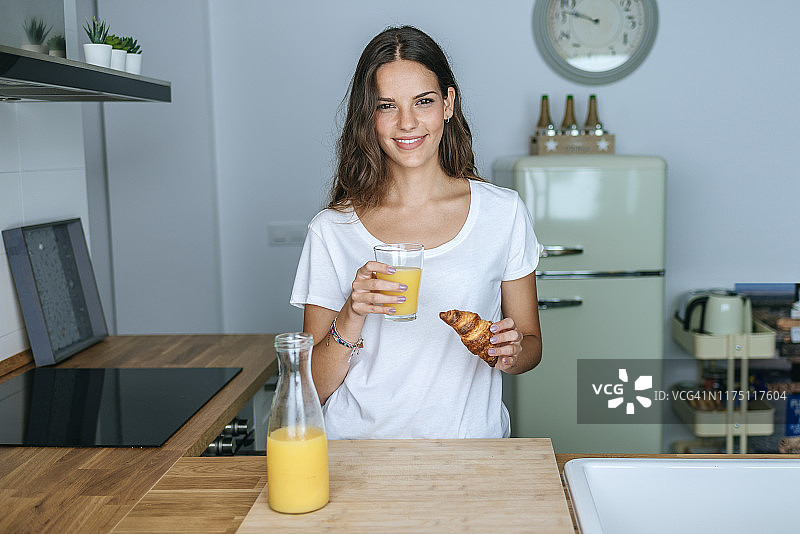 微笑的年轻女子在厨房吃早餐果汁和羊角面包的肖像图片素材