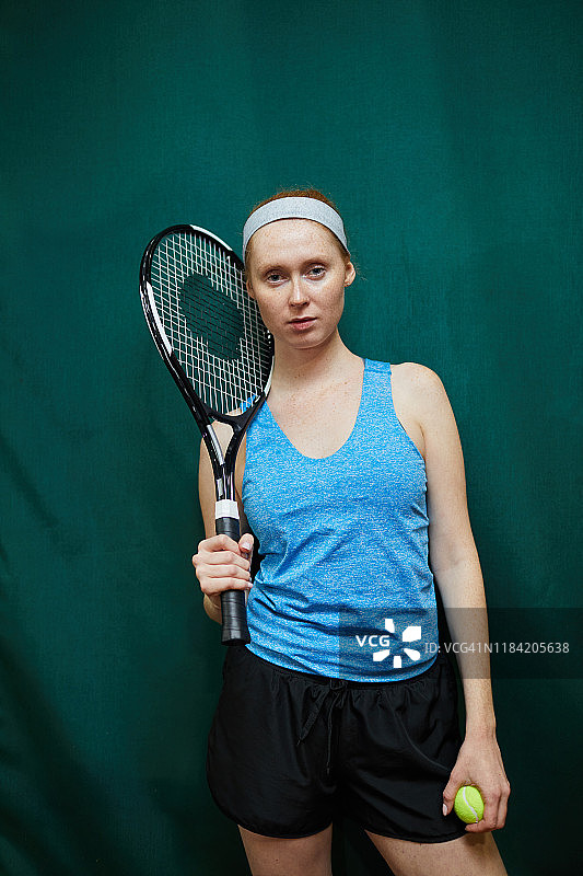 这是一幅严肃自信的年轻女子网球运动员的肖像，她穿着运动服，拿着网球拍和球站在黑暗的背景下图片素材