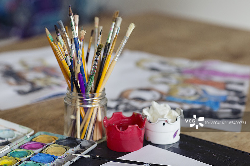 木桌上放着一个装满画笔的玻璃瓶，背景是孩子画的一个家庭图片素材