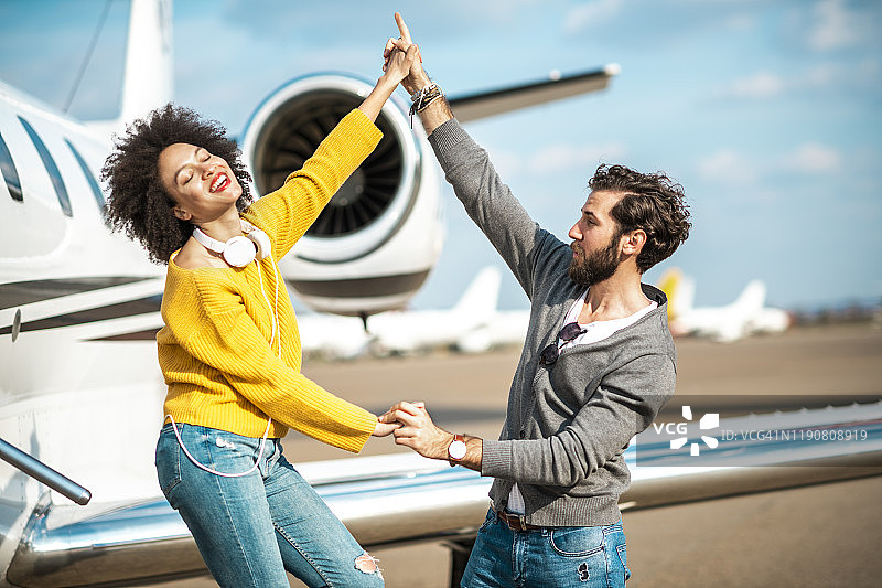 一对年轻时尚的年轻夫妇手牵着手在停机坪上的私人飞机旁跳舞图片素材