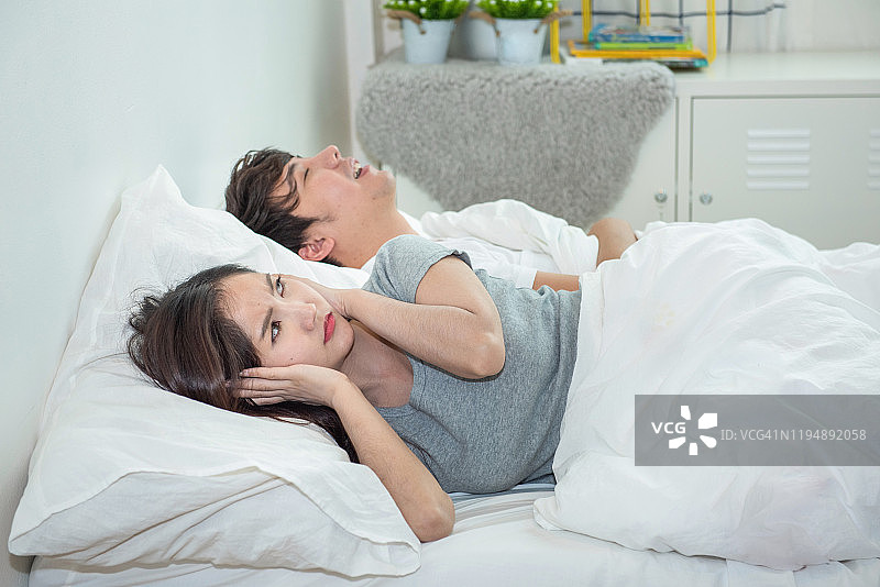 妻子用枕头捂住耳朵时，男人在打呼噜。亚洲年轻女子用枕头捂住耳朵，而她的男友睡在响亮的鼾声中。图片素材