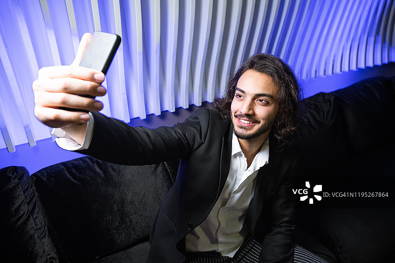 快乐的年轻优雅的男人用智能手机在夜店的沙发上自拍图片素材