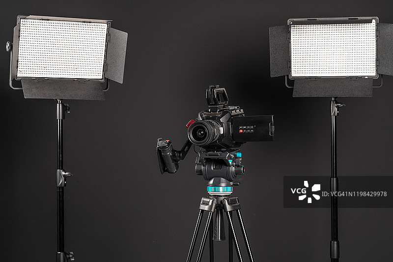 专业影院摄像机在一个摄像机三脚架和一个照明LED面板在黑色工作室背景图片素材