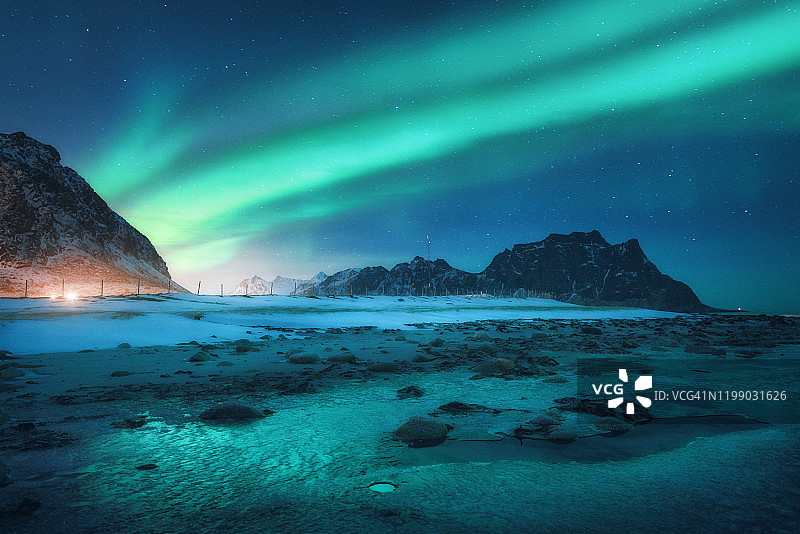 冬天，北极光出现在雪山和沙滩上。挪威罗浮敦群岛的北极光。星空与极地光。夜景有极光，冰冻的海岸，城市的灯光图片素材