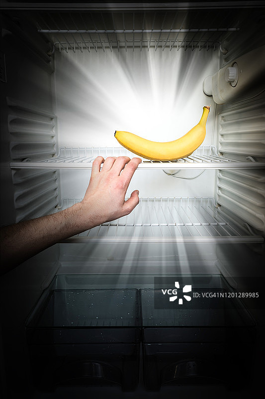 一个男人的手伸向一个在黑暗中发光的香蕉在一个开放的，空的冰箱。减肥饮食概念。图片素材