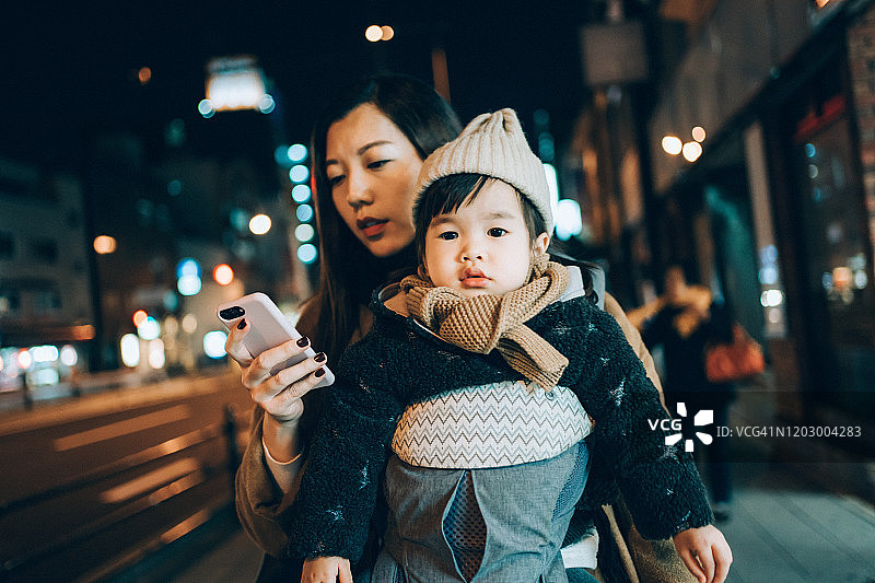 一位年轻的母亲在晚上带着小女儿在城市街道上散步时使用智能手机图片素材