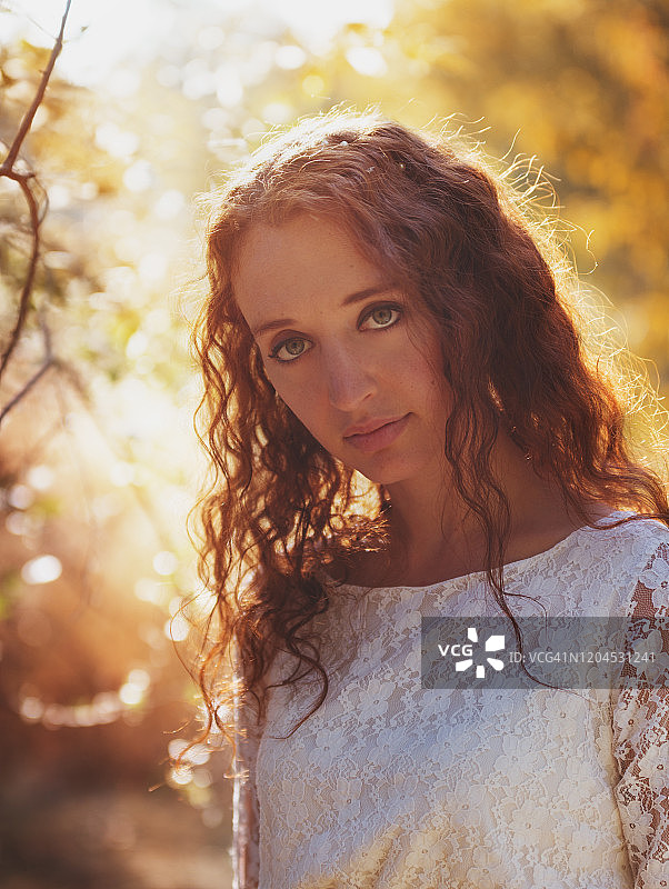 年轻女子的肖像与长长的波浪红头发穿着白色蕾丝连衣裙看着相机与空白的表情在秋天的森林，背光图片素材