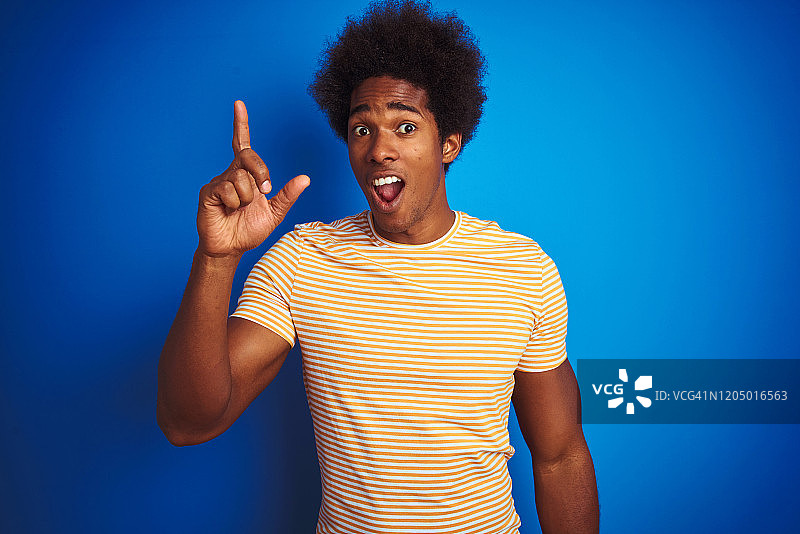 美国男子与非洲式头发穿条纹黄色t恤在孤立的蓝色背景指向手指向上与成功的想法。退出和快乐。第一。图片素材