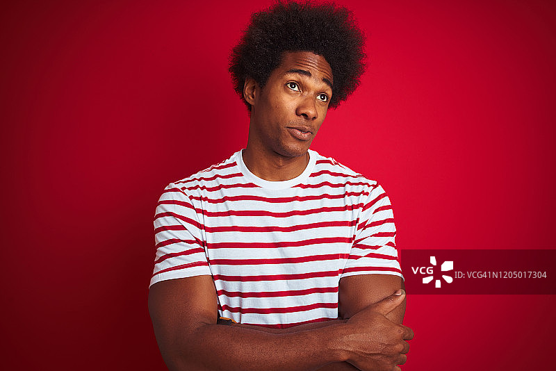 年轻的非洲裔美国人与非洲式头发穿条纹t恤在孤立的红色背景微笑看向一边和凝视远离思考。图片素材