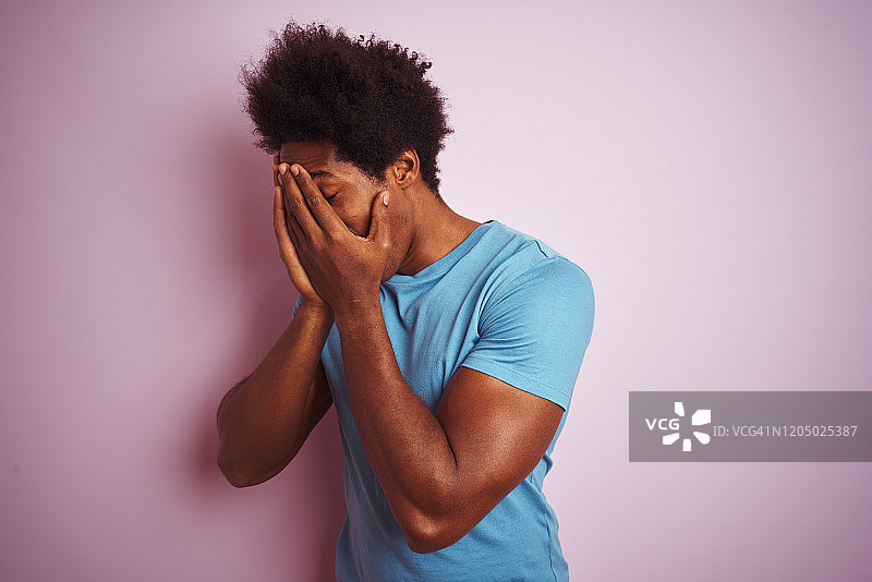 非洲裔美国人与非洲式头发穿蓝色t恤站在孤立的粉红色背景与悲伤的表情覆盖脸与手哭泣。抑郁症的概念。图片素材