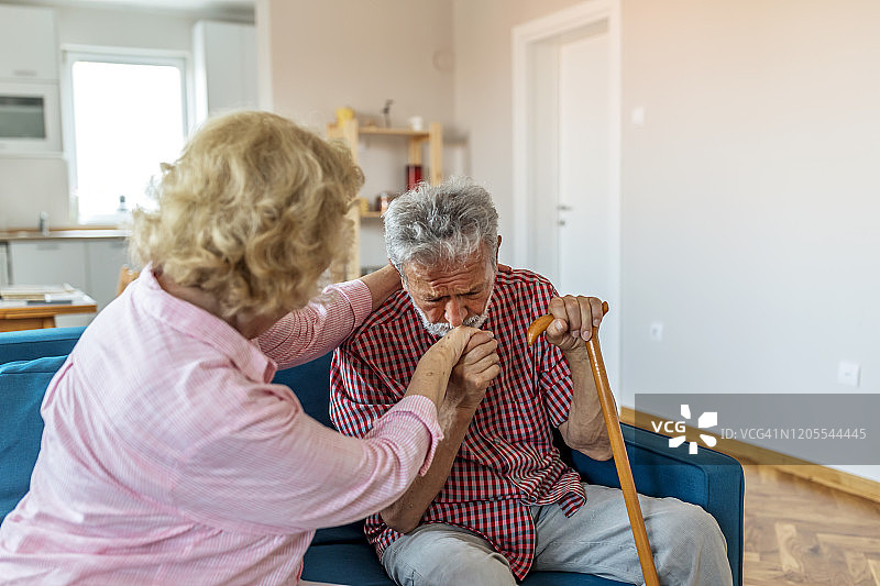 悲伤的退休丈夫拄着拐杖坐在客厅里，旁边是老妻子。老夫妇看着对方，手牵着手在家里图片素材