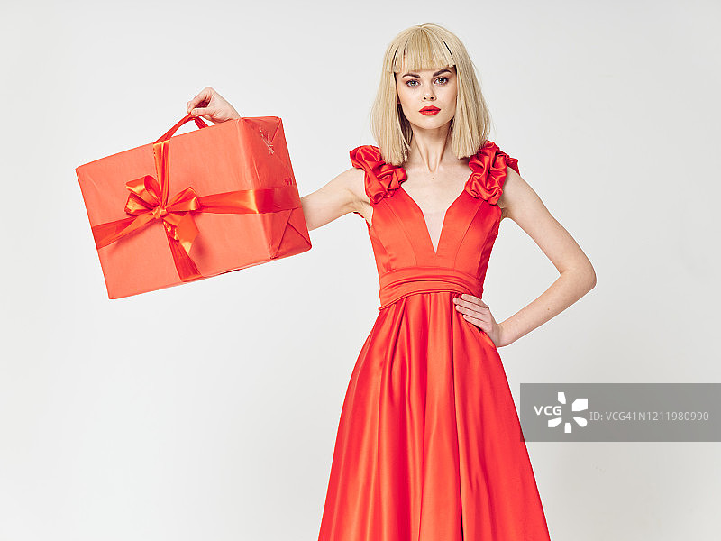 一个穿着红色节日礼服的年轻女孩手里拿着一个装着礼物的大红色盒子。图片素材