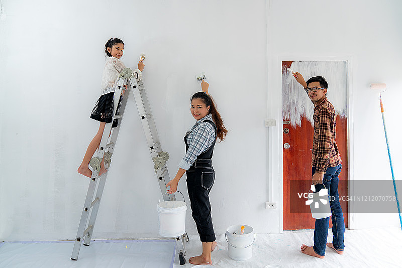 幸福的亚洲家庭与父亲，母亲和女儿在粉刷粉刷白色的墙壁在他们的新房子。家庭生活爱情关系，还是新房活动概念图片素材