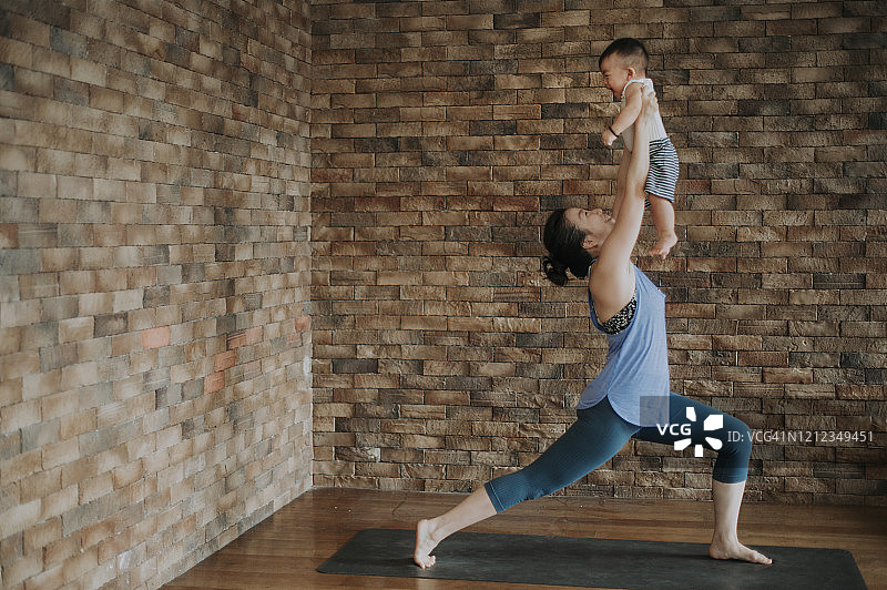 一位亚裔中国女瑜伽教练和她9个月大的儿子一起在客厅的砖墙上练瑜伽图片素材