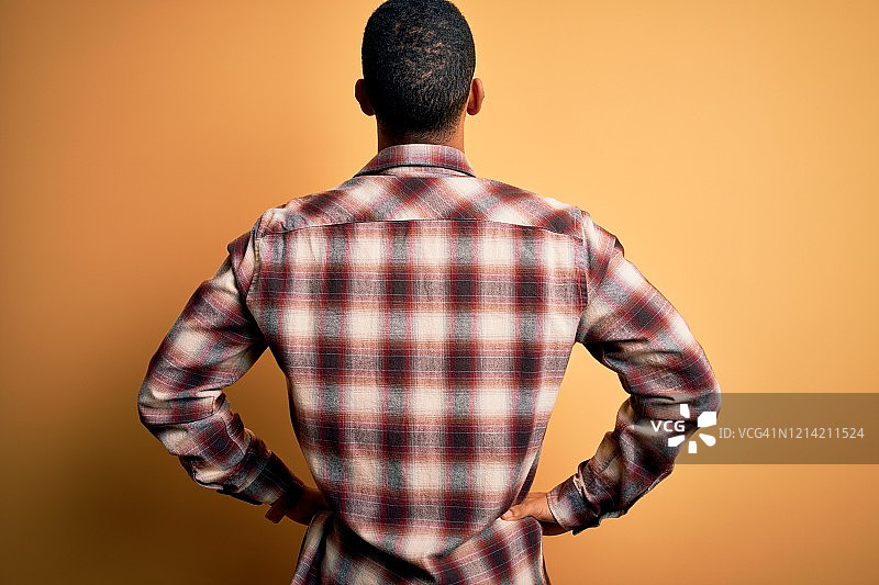 年轻英俊的非裔美国人穿着休闲衬衫站在黄色背景站向后看离开与手臂上的身体图片素材
