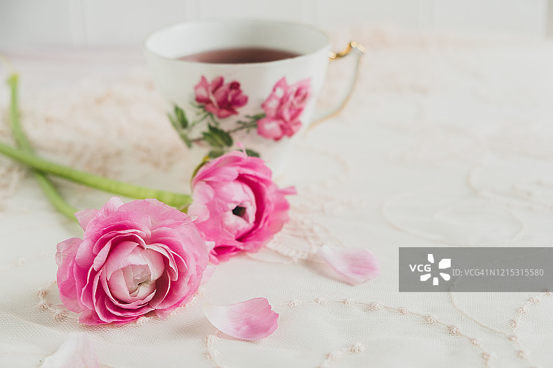 粉彩风格的股票形象的粉红色毛茛花在一个花边覆盖的桌面与一杯茶在背景图片素材