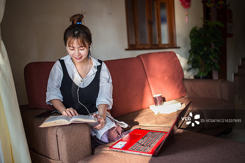 一位亚洲华裔年轻女性在家工作。女商人坐在沙发上用笔记本电脑工作。年轻女子喜欢在工作期间喝咖啡。她在休息时间读书。图片素材