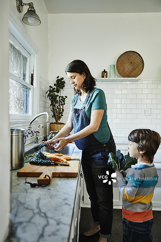 怀孕的母亲在厨房的水槽里洗蔬菜，小儿子在一旁看着图片素材