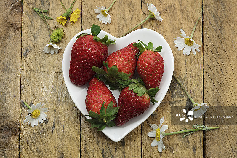 新鲜的熟透的有机草莓放在一个心形瓷碗里，中间点缀着野花，乡村的木质背景和复制空间。图片素材