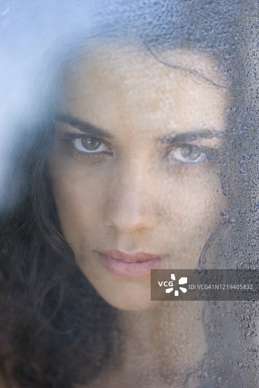 近距离拍摄的黑发女人透过满是雨滴的窗户往外看图片素材