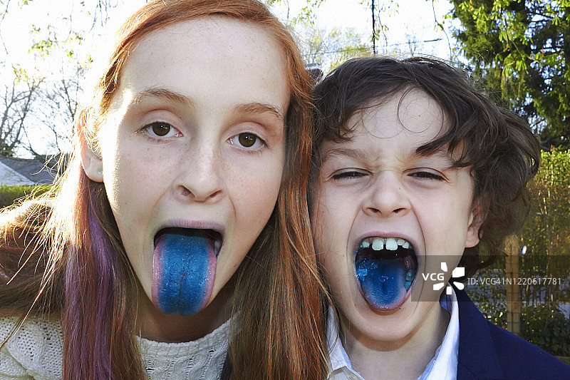 一个红头发的妹妹和一个留着棕色短卷发的弟弟对着镜头伸着舌头。因为吃了什么，它们的舌头都是亮蓝色的。可爱,有趣的图片。他们在外面，后面有树。图片素材
