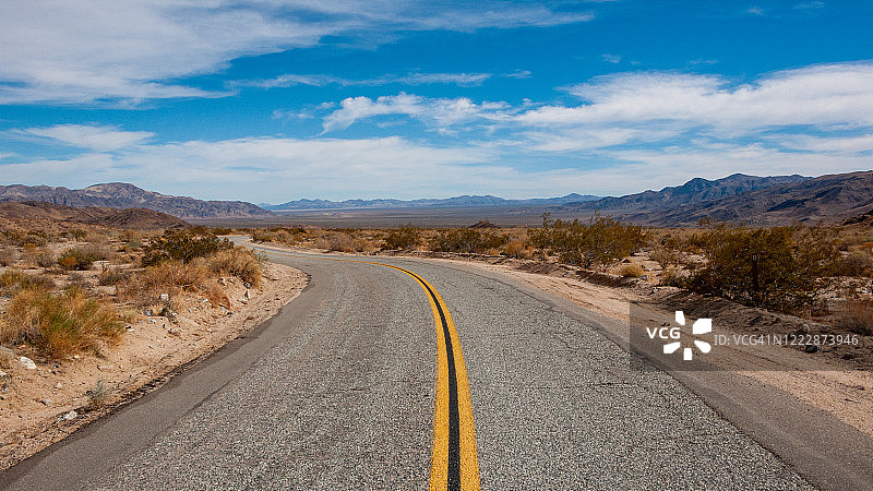 加州沙漠的空旷道路，中间有一条双黄线，蓝天上有稀疏的云彩。道路转弯进入远方的消失点图片素材