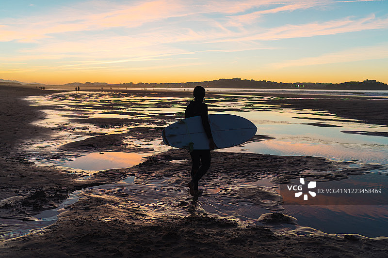 日落时分，年轻的男子冲浪者腋下夹着冲浪板走在沙滩上图片素材