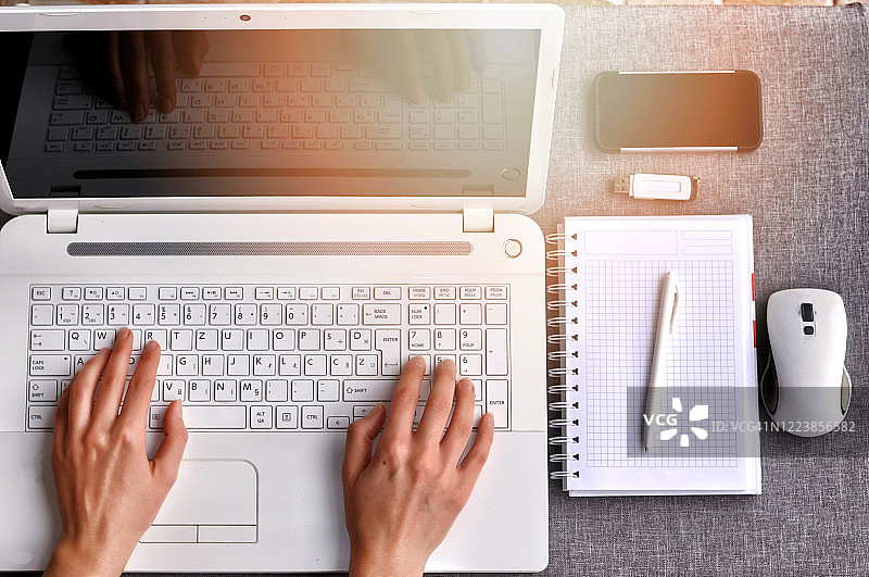 人类的双手在一个白色的笔记本电脑键盘上打字，屏幕是隔离的。工作台工作台概念。上面的办公室工作场所与手机和鼠标，笔记本，笔和usb闪存在灰色的桌子上。图片素材